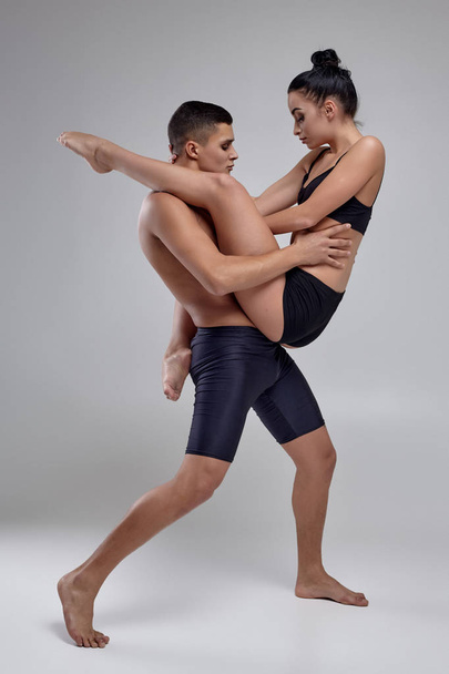 Το ζευγάρι ενός νεαρού σύγχρονου χορού μπαλέτου με μαύρα κοστούμια ποζάρει σε ένα γκρίζο φόντο στούντιο. - Φωτογραφία, εικόνα