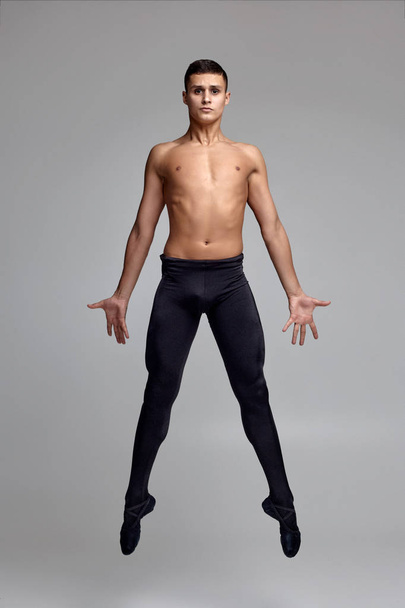 Photo of an athletic man ballet dancer, dressed in a black tights and pointe, making a dance element against a gray background in studio. - Foto, Imagen
