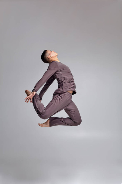 Photo of an athletic man ballet dancer dressed in a gray tracksuit, making a dance element against a gray background in studio. - Photo, image