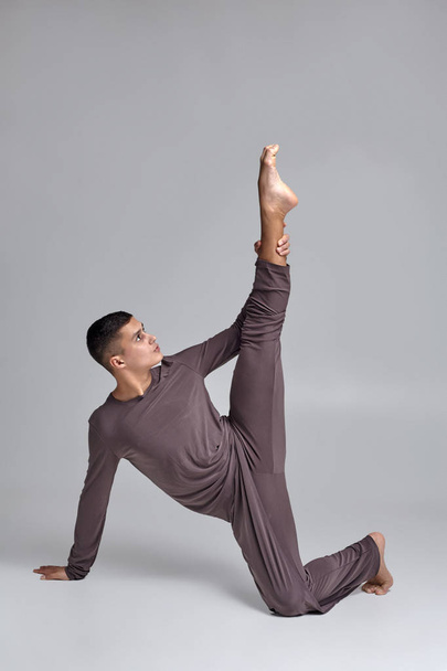Photo of an athletic man ballet dancer dressed in a gray tracksuit, making a dance element against a gray background in studio. - Photo, Image