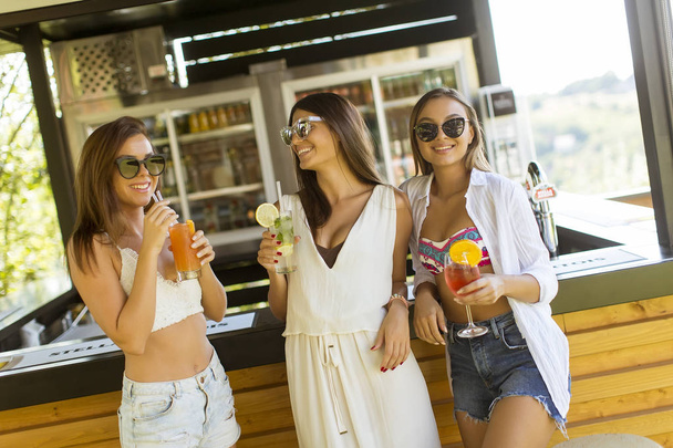 Τρεις όμορφες νεαρές γυναίκες που πίνουν κοκτέιλ και διασκεδάζουν στο Beach Bar - Φωτογραφία, εικόνα