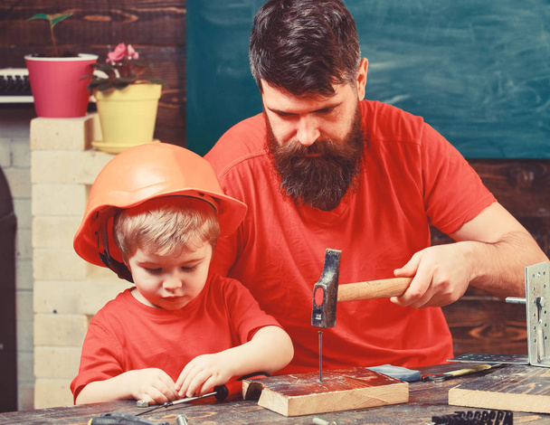 Boy, child busy in protective helmet learning to hammering hobnails with dad. Father, parent with beard teaching little son to use hobnails and hammer. Workshop and handyman concept - Фото, изображение