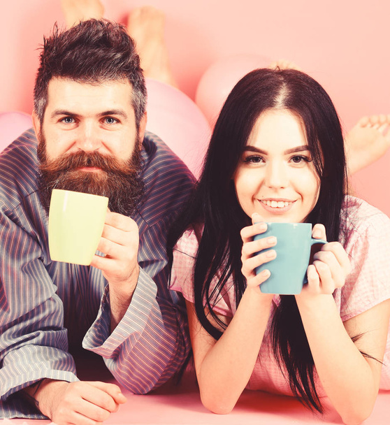 Couple relax in morning with coffee. Family tradition concept. Man and woman on smiling faces lay, pink background. Couple in love drink coffee in bed. Man and woman in domestic clothes, pajamas - Photo, image