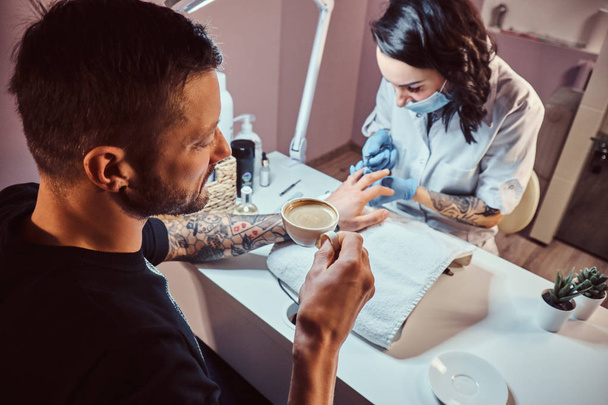 Een man het ontvangen van een manicure in de schoonheidssalon, ontspannen en drinken van een kopje koffie, terwijl schoonheidsspecialiste Master doet manicure procedure - Foto, afbeelding