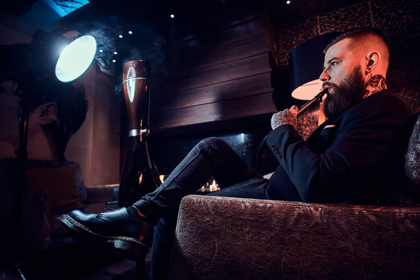 Hombre guapo tatuado se relaja en el sillón y está fumando narguile
 - Foto, Imagen