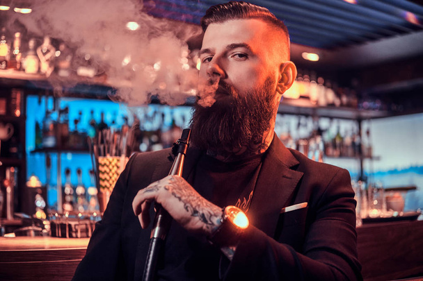 Retrato de hombre barbudo tatuado que está fumando narguile, haciendo buen vapor
 - Foto, imagen