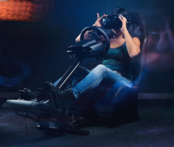 Γυναίκα φορώντας ακουστικά Vr οδήγηση σε αυτοκίνητο αγωνιστικά πιλοτήριο προσομοιωτή με κάθισμα και τροχό. - Φωτογραφία, εικόνα