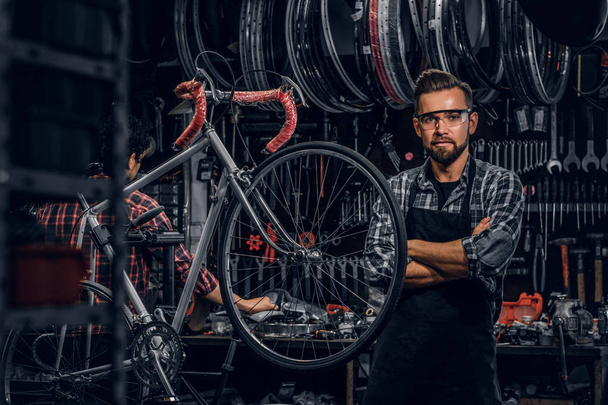 Knappe bebaarde man in glazen staat in de buurt van vaste fiets in zijn eigen workshop - Foto, afbeelding