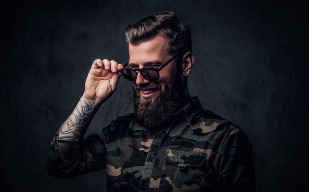 Retrato de un elegante barbudo con las manos tatuadas en la camisa militar tomados de la mano en gafas de sol y se ríe. Estudio foto contra la pared oscura
 - Foto, imagen