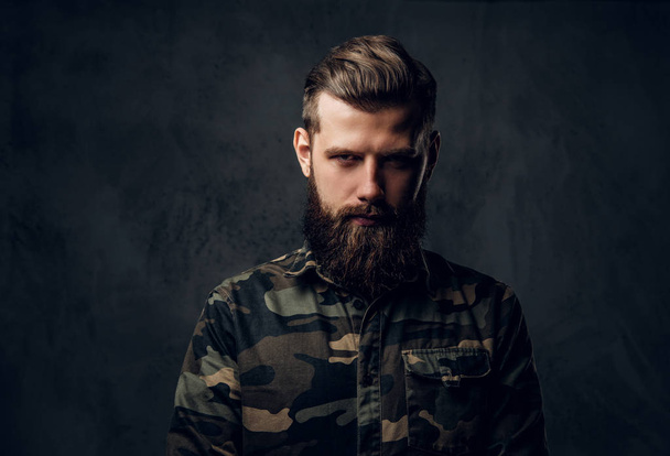 Portrait of a stylish bearded guy with tattooed hands in the military shirt. Studio photo against dark wall - Photo, Image