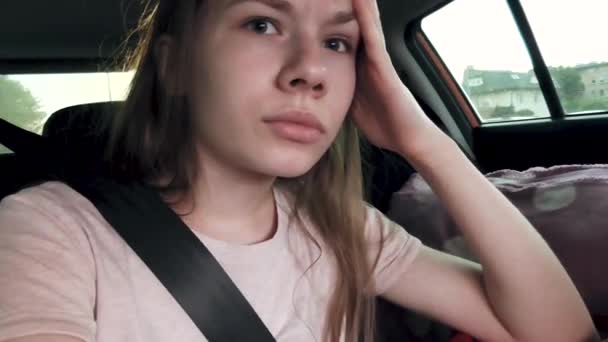The girl is sitting in the back seat of the car, wearing a seatbelt and resenting about something, thinking and looking around, moving her eyebrows and lips. The car does not move. - Footage, Video