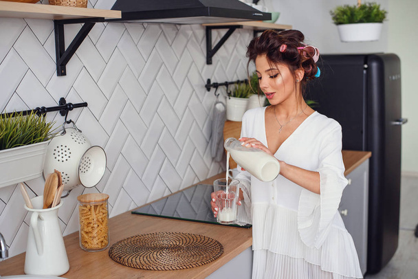 Beautiful girl dressed in white bathrobe pours milk from a carafe into a glass, in the stylish cozy kitchen. Healthy eating and diet concept. - Zdjęcie, obraz