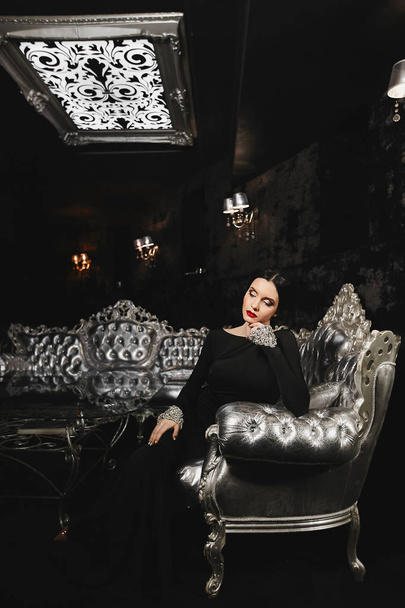 Fashionable and sexy brunette model woman with perfect body and with red lips in black evening dress with sits on the silver sofa at modern luxury interior - Fotoğraf, Görsel
