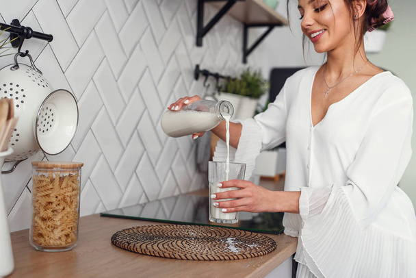 Beautiful smiling woman pours fresh milk from a carafe into a glass, in the stylish cozy kitchen at the morning. Healthy eating and diet concept. - 写真・画像