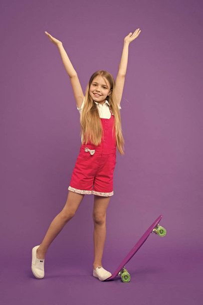 Childhood and active games. Small girl smile with skate board on violet background. Carefree day. Child skater smiling with longboard. Skateboard kid in pink jumpsuit. Sport activity and energy - Photo, image