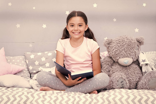 Read fairytale before go to bed. Girl child sit bed with teddy bear read book. Kid prepare to go to bed. Pleasant time in cozy bedroom. Girl kid long hair cute pajamas relax and read book to bear toy - 写真・画像