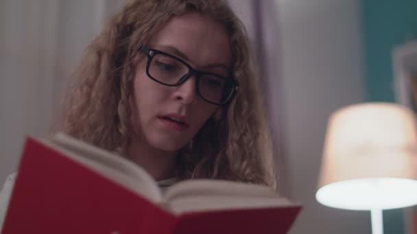 Young beautiful woman reads a paper book in her cozy living room in the evening - Materiaali, video