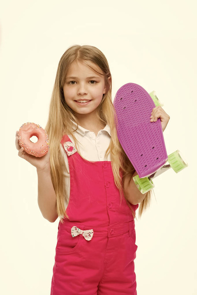 Meisje glimlach met skateboard en donut geïsoleerd op wit. De schaatser van het kind met de donut en longboard. Skateboard kid met junk food. Sport en energie. Voedsel voor snack en dessert. dieet concept - Foto, afbeelding