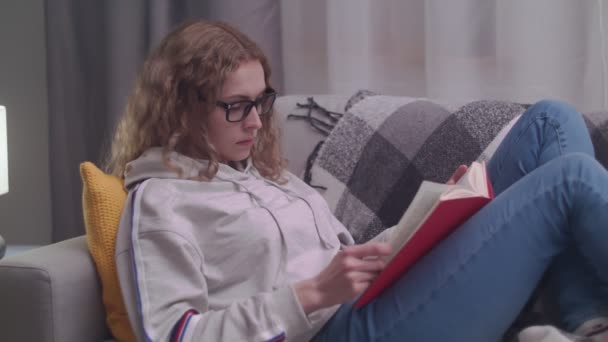 Young beautiful woman reads a paper book in her cozy living room in the evening - Filmmaterial, Video