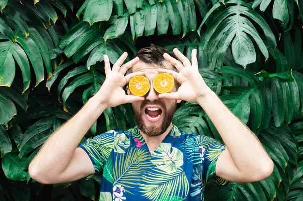Young bearded man holding slices of orange tangerine in front of his eyes, laughing - Photo, image