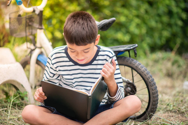 Pieni poika lukemassa kirjaa istuu polkupyörän puistossa
 - Valokuva, kuva