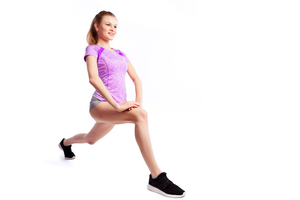 Een jonge vrouw coach in een sportieve korte top en Gym legging maakt longen door de voeten naar voren, handen worden gehouden aan de zijkant op een witte geïsoleerde achtergrond in de Studio  - Foto, afbeelding