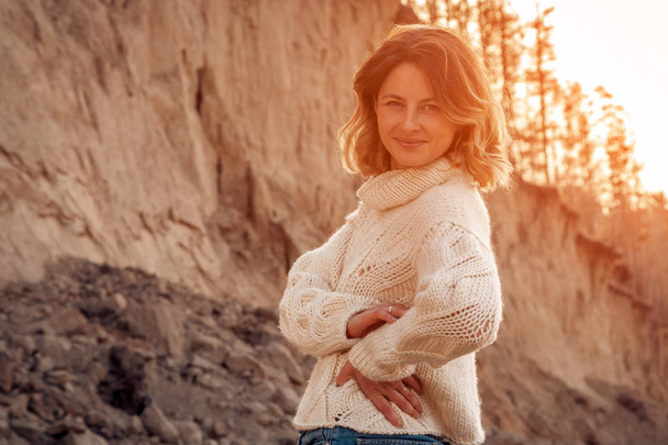 Retrato de una hermosa modelo joven en ropa de abrigo disfrutar del día en la playa; en el fondo el mar con puesta de sol. Foto cálida de otoño. Mujer sonriendo y mirando hacia otro lado
 - Foto, Imagen