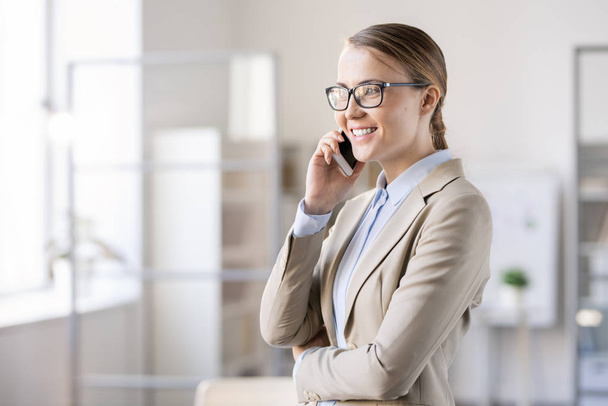 Heureuse jeune femme gestionnaire confiante dans des lunettes debout dans le bureau et parler au client au téléphone tout en discutant bonne affaire
 - Photo, image