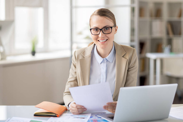 Cheerful confident attractive financial employee in glasses sitting at desk with diary and laptop and examining report, she looking at camera - Photo, Image