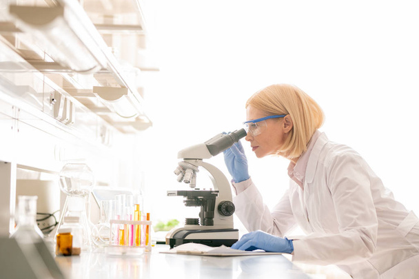 Grave ricercatore scientifico maturo impegnato con capelli biondi in piedi al banco di laboratorio e campione di studio attraverso il microscopio
 - Foto, immagini