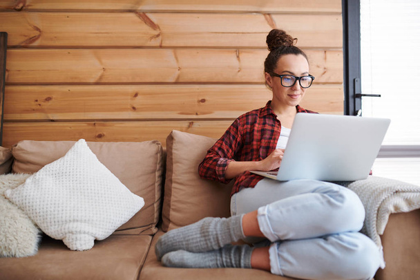 Jovem freelancer feminino casual sentado no sofá com laptop na frente enquanto trabalhava na rede em casa
 - Foto, Imagem