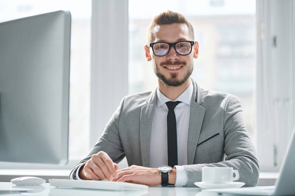 Happy young successful businessman in eyeglasses and suit sitting by desk in front of computer monitor - Foto, Imagem