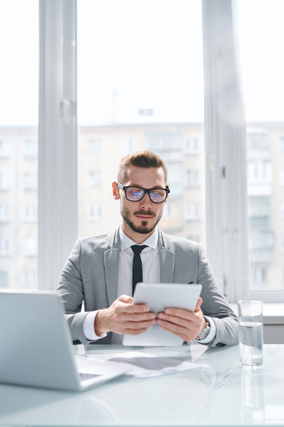 Młody poważny ekonomista w formalnym ubraniu koncentrujący się na czytaniu danych online w tabletkach podczas pracy w biurze - Zdjęcie, obraz