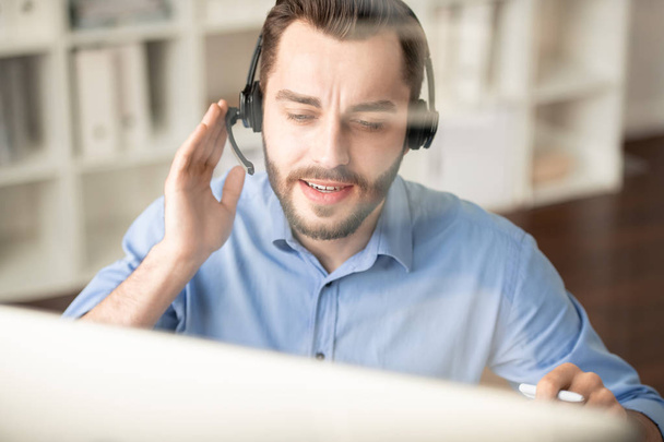 junger selbstbewusster Manager mit Kopfhörer, der auf den Computerbildschirm blickt, während er Kunden berät und ihre Fragen beantwortet - Foto, Bild