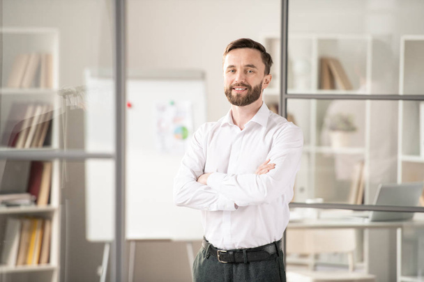 Young bearded office worker crossing his arms on chest while standing in front of camera on background of workplace - Foto, Bild