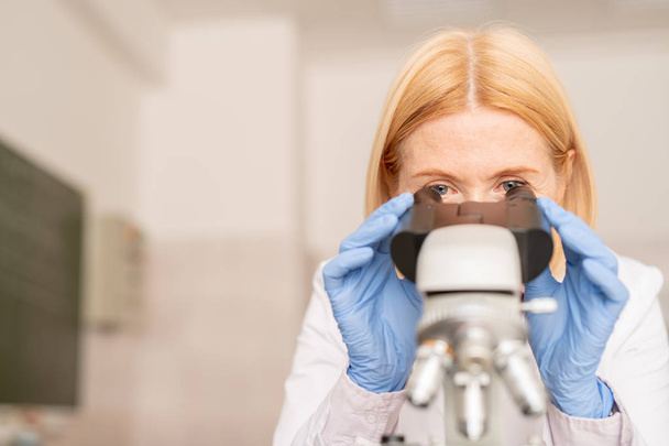 ernste konzentrierte blondhaarige Laborspezialist in sterilen Handschuhen unter dem Mikroskop während der wissenschaftlichen Forschung - Foto, Bild