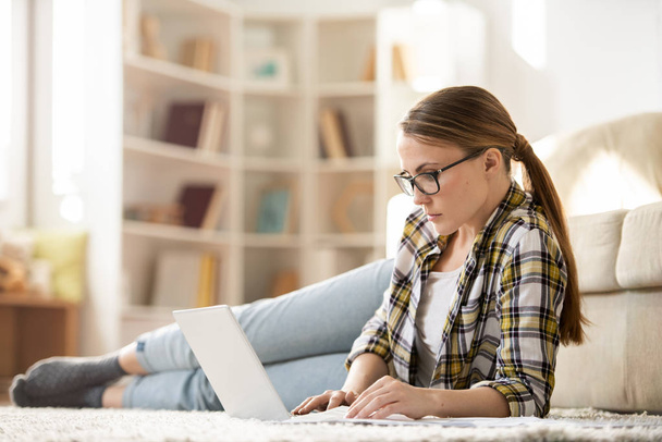 自宅で仕事をしながら、カーペットの上に横たわってノートパソコンを使用してポニーテールを持つ深刻なペンシブ若い女性フリーランサー - 写真・画像