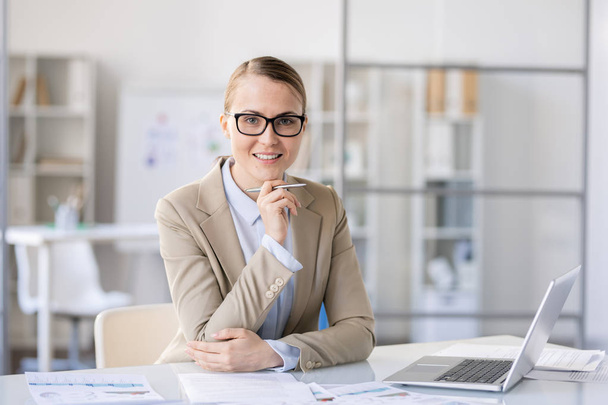 Cheerful successful female business researcher in jacket sitting at desk in office and holding pen while looking at camera confidently - Fotoğraf, Görsel