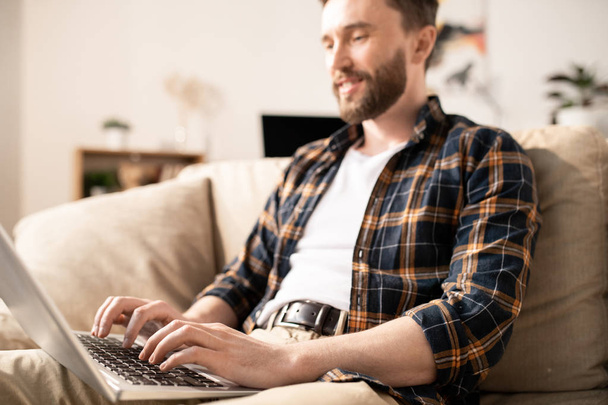 Joven hombre de negocios móvil en ropa casual escribiendo en el teclado del ordenador portátil mientras mira la pantalla durante la red en el sofá
 - Foto, Imagen
