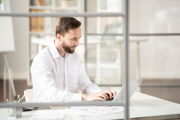 Зайнятий молодий працівник у білій сорочці, що друкує на клавіатурі ноутбука, дивлячись на дисплей під час роботи в офісі
 - Фото, зображення