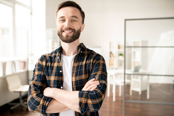 Homem casual jovem barbudo com sorriso de dente olhando para você na pausa no ambiente de escritório enquanto desfruta de descanso
 - Foto, Imagem