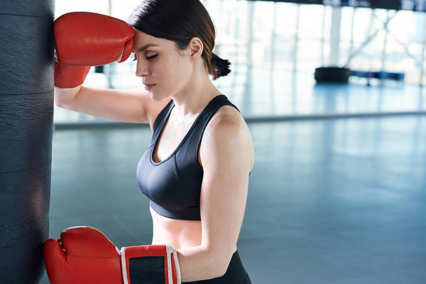 Young tired brunette female boxer standing by punch bag in gym while having short rest before new training - Φωτογραφία, εικόνα