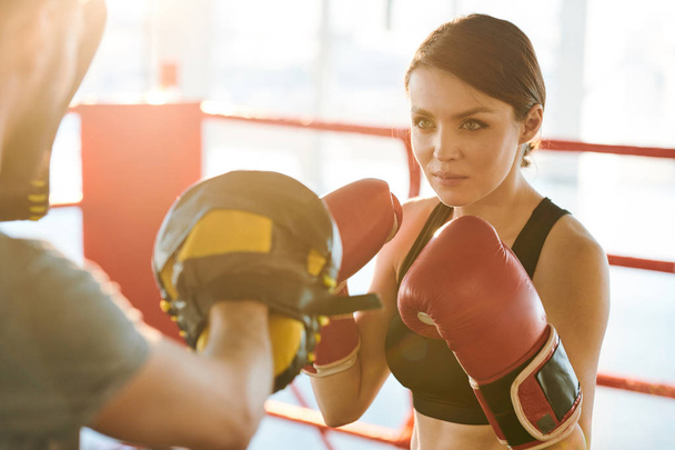Młody poważny bokser kobiet patrząc na jej rywala gotowy do ataku go podczas treningu na pierścieniu bokserskim - Zdjęcie, obraz