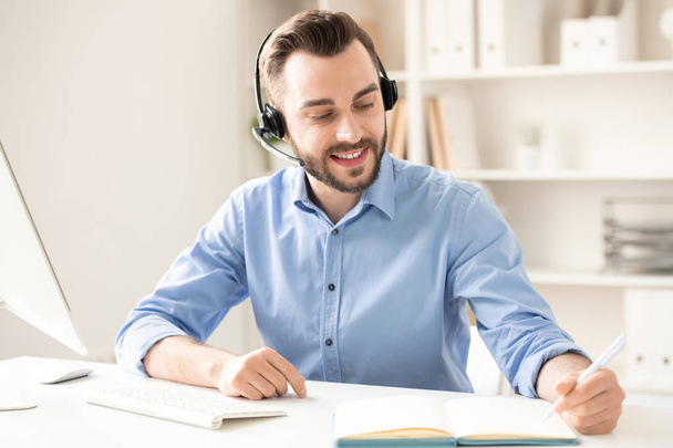 Ευτυχισμένος νέος άντρας με ακουστικά που κάθονται από τη ρεσεψιόν, συμβουλευτούν τους πελάτες και κάνοντας σημειώσεις εργασίας στο σημειωματάριο - Φωτογραφία, εικόνα