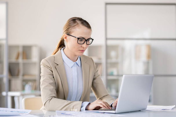 Modern ofiste masada oturan ve dizüstü bilgisayarda sunum hazırlarken klavyede yazarak gözlük ciddi meşgul genç kadın - Fotoğraf, Görsel