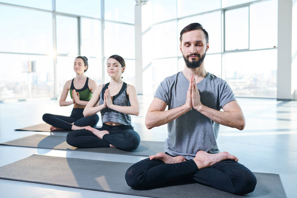 Bearded young man with crossed legs and hands put together by chest sitting on mat on background of two exercising girls - Foto, Bild