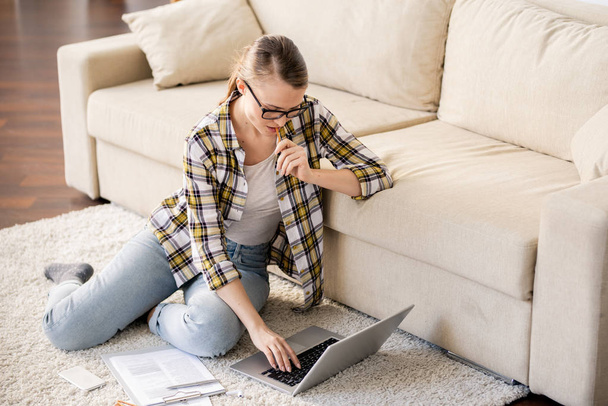Серьезный молодой внештатный агент, сидящий на полу и использующий ноутбук во время работы с бумагами, она думает о докладе дома
 - Фото, изображение