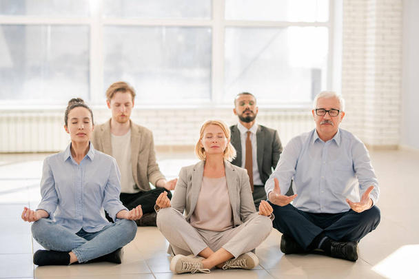 Group of young and mature people in formalwear sitting on the floor in pose of lotus during meditation - Foto, afbeelding