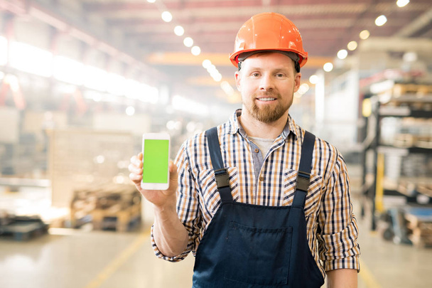 Young successful factory engineer in overalls and helmet showing promo on touchscreen of his smartphone - Photo, Image