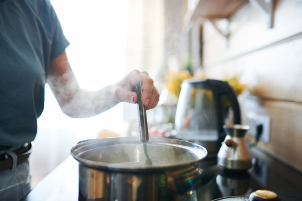 Hand van jonge huisvrouw met lepel in pan tijdens het mengen van warm eten in de keuken - Foto, afbeelding